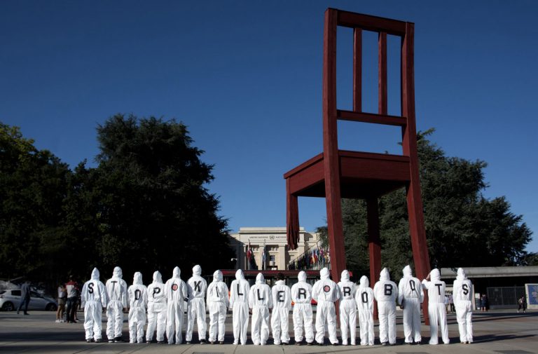 activistas de pie en una fila debajo de la escultura de la Silla Rota fuera de la ONU en Ginebra. Los activistas llevan trajes de caldera con letras en la parte posterior para deletrear