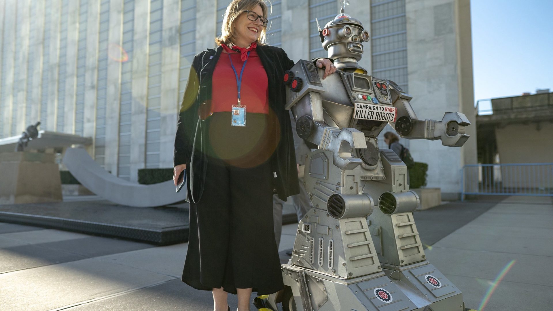 Mary Wareham poses with her hand on the Campaign robot’s shoulder in front of the UN building with the sun behind them.