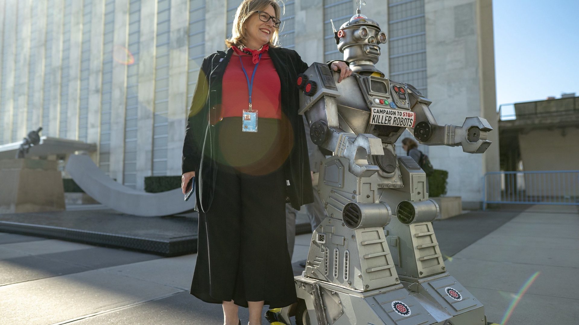 Mary Wareham posa con la mano en el hombro del robot de la Campaña frente al edificio de la ONU con el sol detrás de ellos.