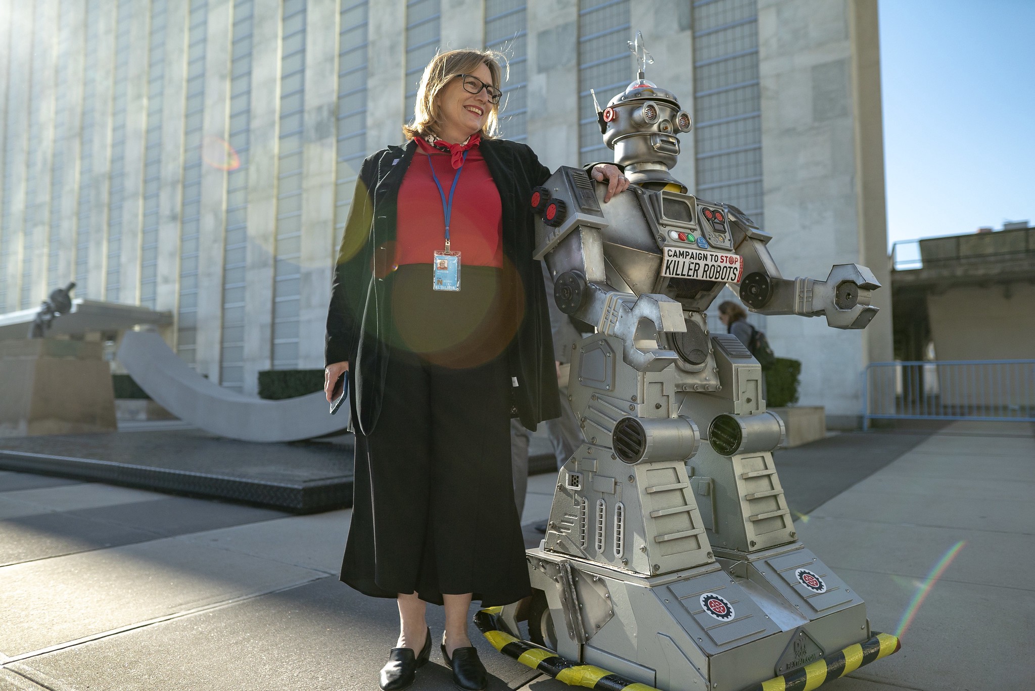 Mary Wareham poses with her hand on the Campaign robot’s shoulder in front of the UN building with the sun behind them.
