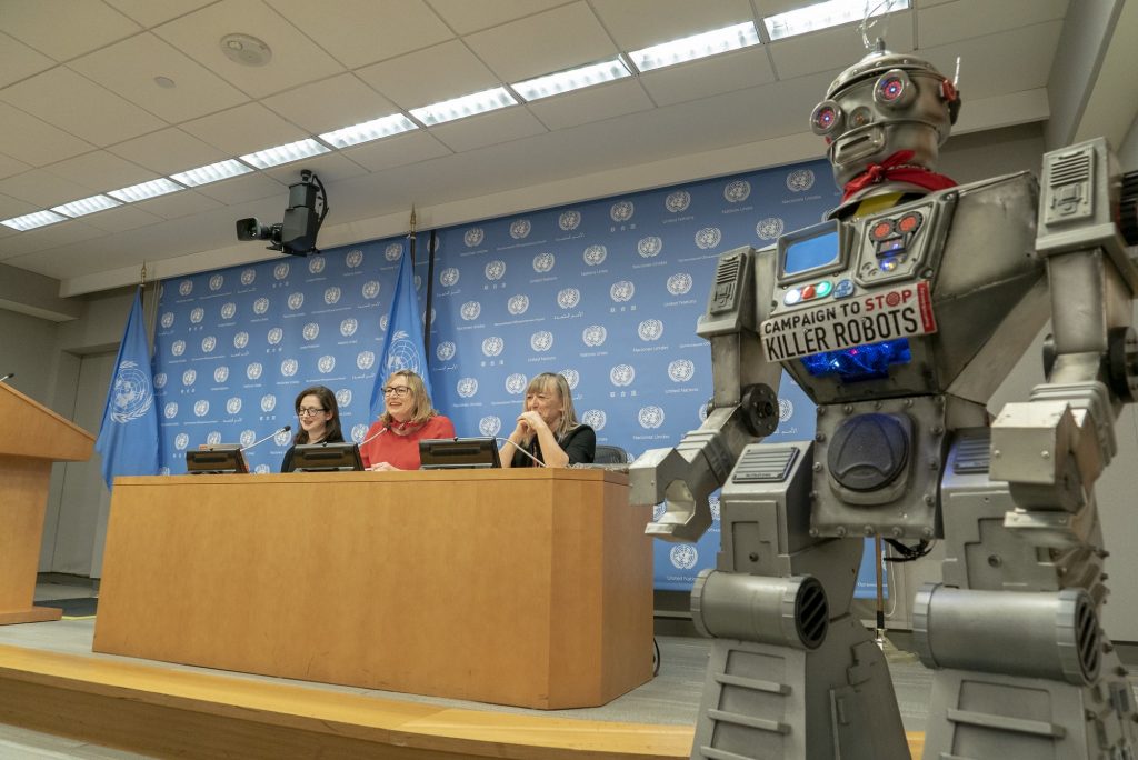 Liz O'Sullivan, Mary Wareham y Jody Williams sonríen a los periodistas desde su escritorio durante una conferencia de prensa, el robot de la Campaña está a la derecha de la escena.