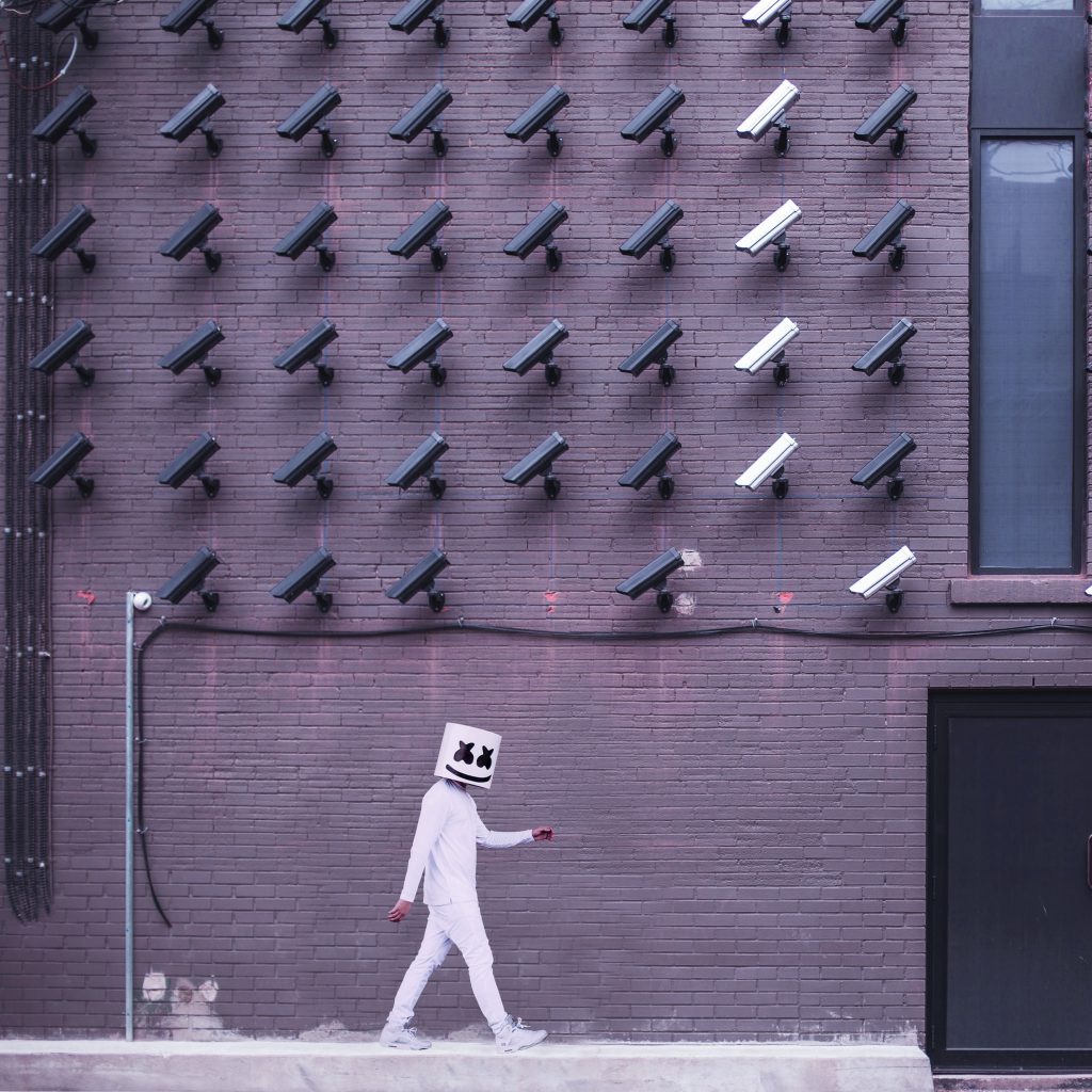 CCTV cameras fill a brick wall pointing at the footpath while a black man in white clothing, with a head covering that has a smiley face with ‘X’ eyes walks by.