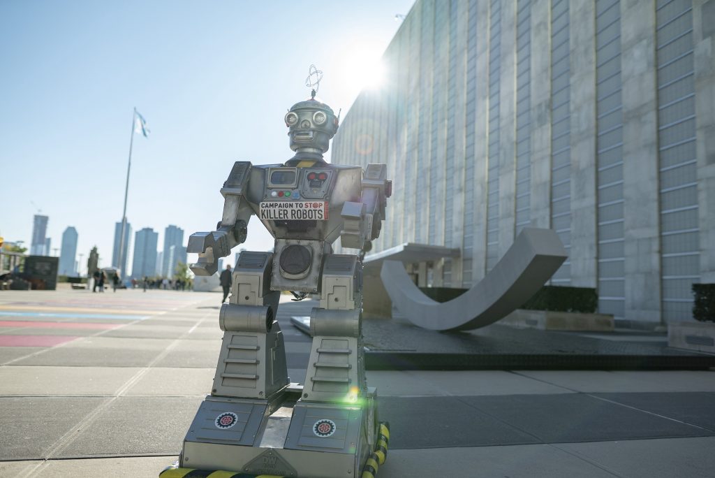 El robot Campaign to Stop Killer Robots se encuentra fuera del edificio de la ONU en Nueva York mientras el sol asoma sobre el edificio. La bandera de la ONU ondea en la distancia.
