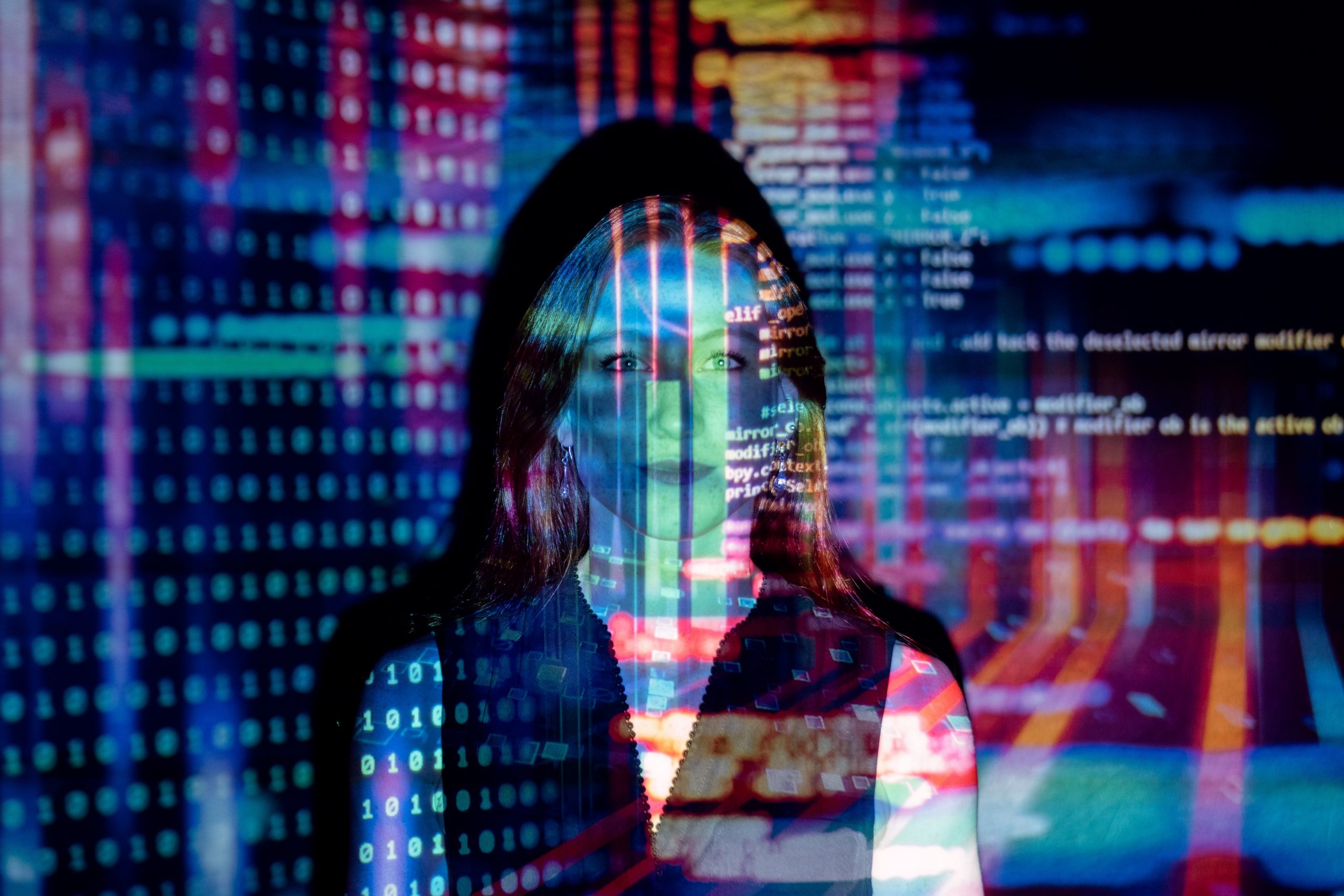 A female software engineer looks directly at the camera with colourful code projected on her.